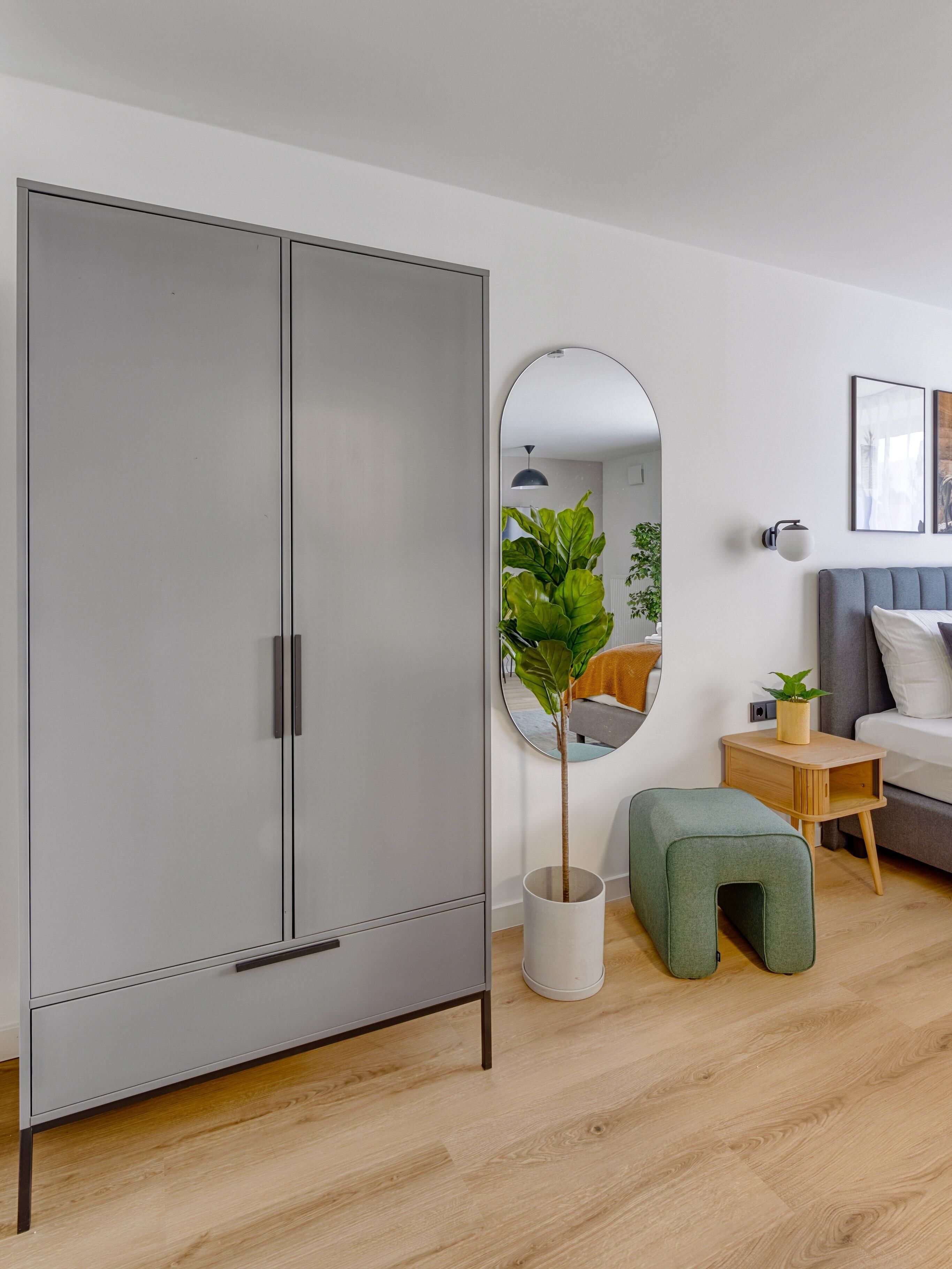 Un dormitorio moderno con un armario gris, un espejo redondo, un reposapiés verde, una mesita de noche de madera y un sofá gris.