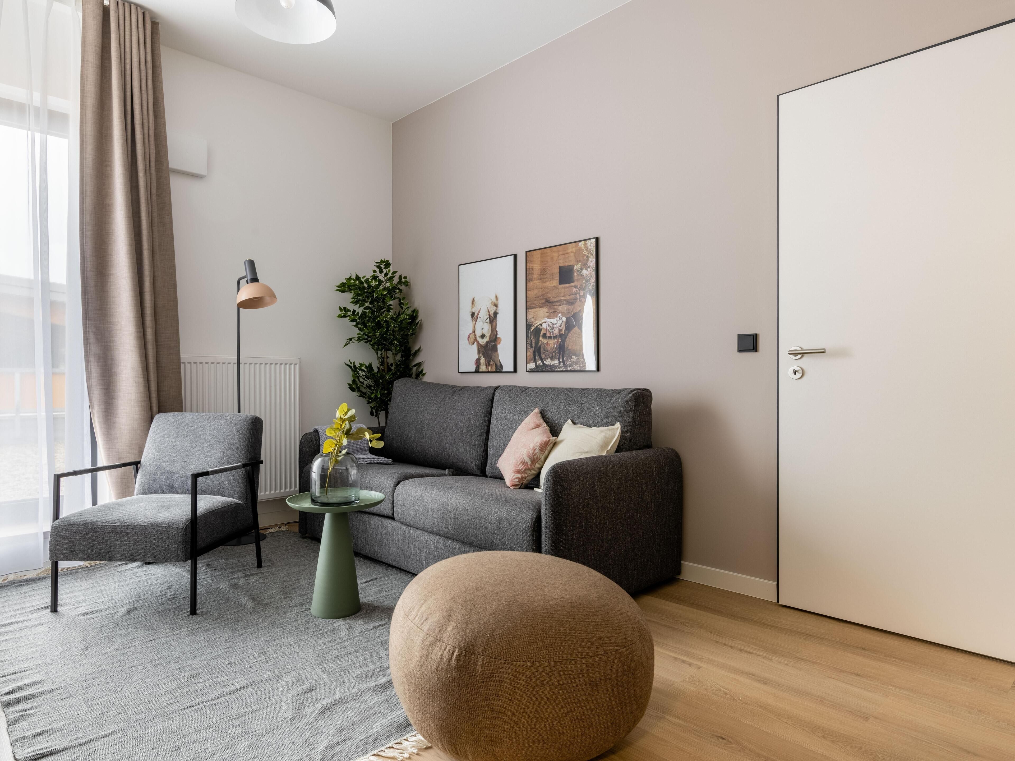 Un salon moderne avec un canapé gris, un pouf rond, une table d’appoint verte et un lampadaire. Deux tableaux encadrés ornent le mur, ainsi qu’une plante en pot et une fenêtre avec des rideaux.