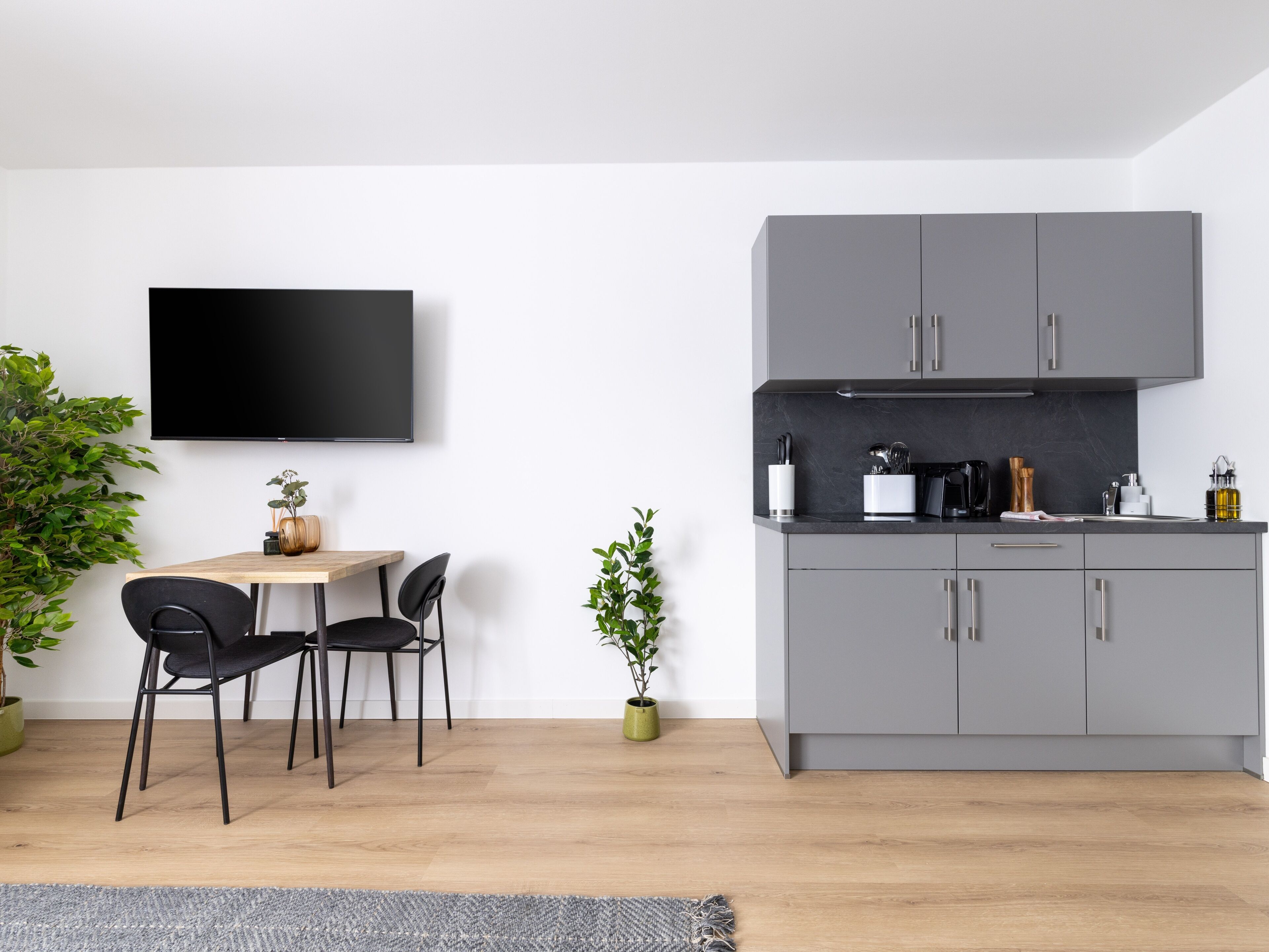 A modern kitchen with grey cabinets, a flat-screen TV, and a dining area with two chairs.