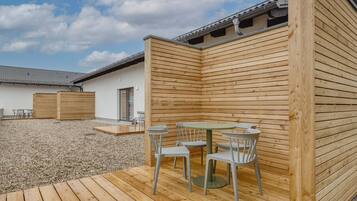 Une terrasse en bois avec une table et des chaises, un espace extérieur aménagé et couvert, et un bâtiment doté d’un toit en tuiles.