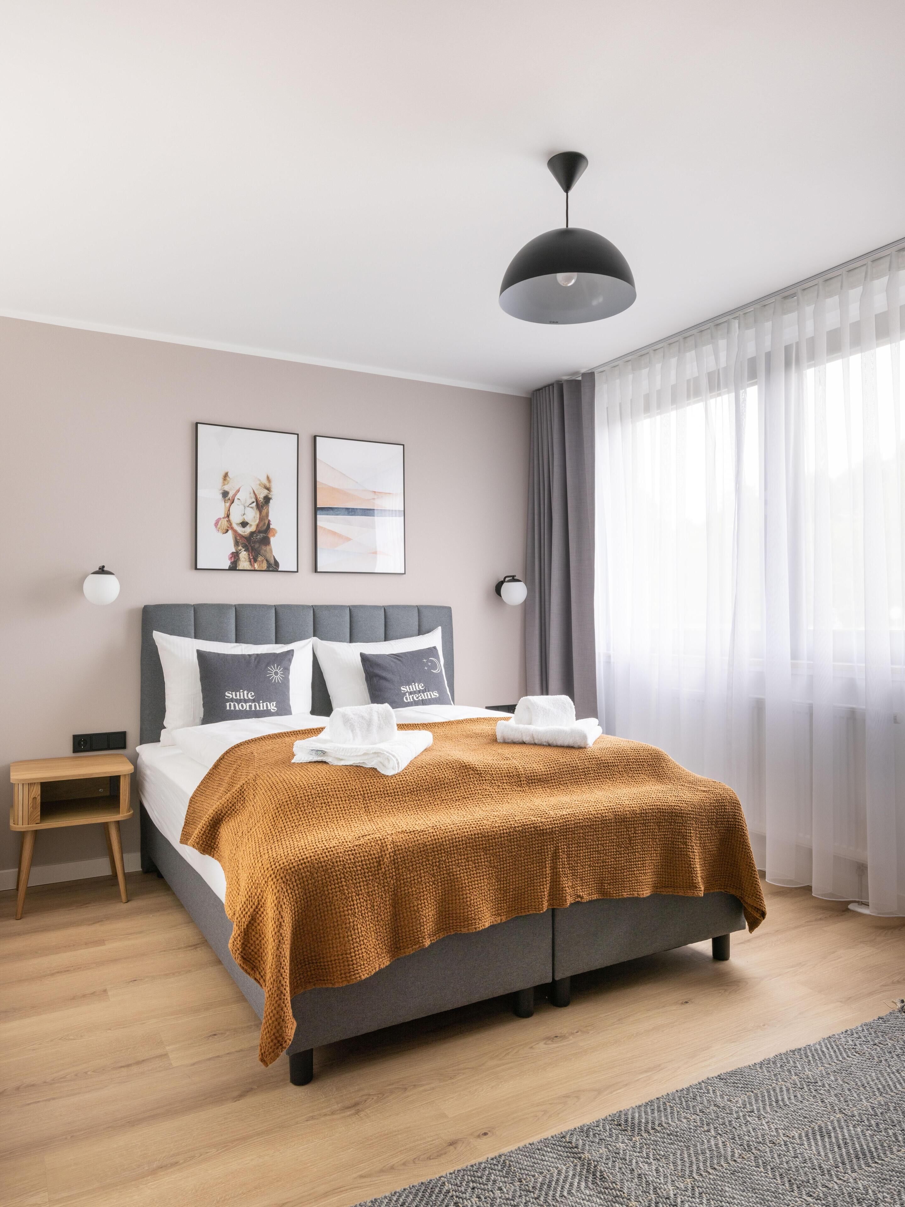 Un dormitorio moderno con una cama gris, un edredón marrón, dos almohadas con texto, una mesita de noche pequeña y un ventanal con cortinas traslúcidas.