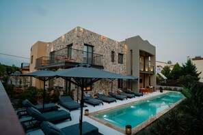 A modern building with a stone facade, a swimming pool, and outdoor seating with umbrellas. - Capital Luxury Suites (Nafplio)