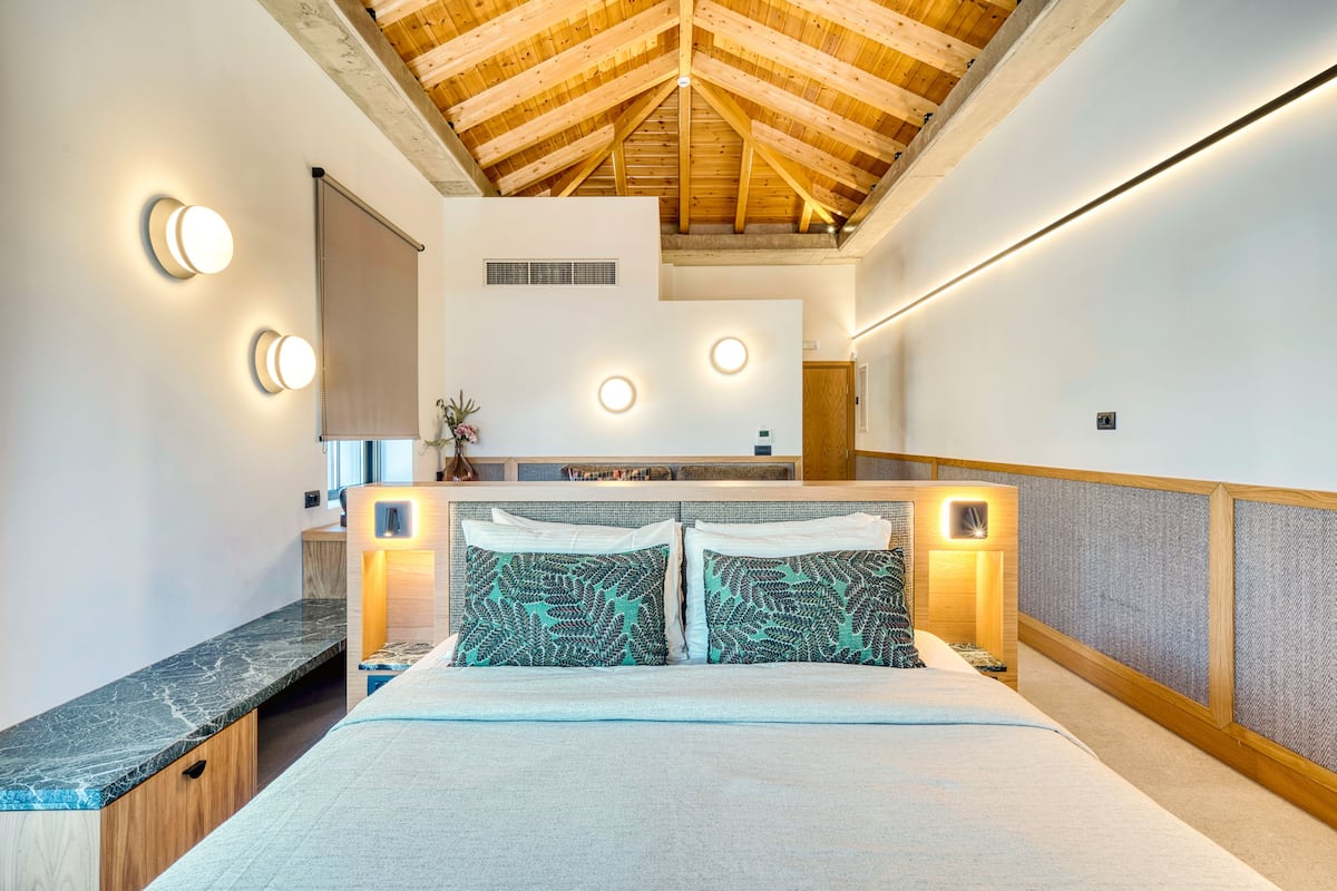Un dormitorio moderno con techo alto, cama con almohadas estampadas, cabecera de madera e iluminación empotrada.