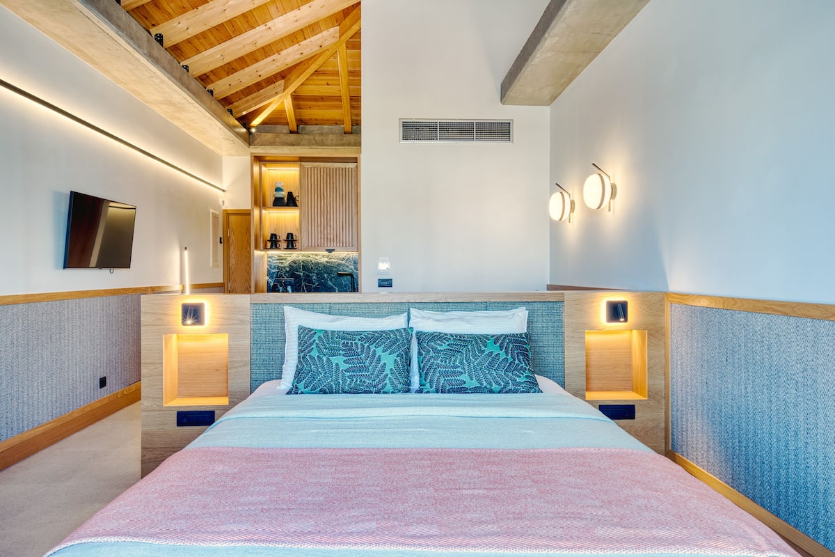 Un dormitorio moderno con una cama grande, vigas de techo de madera, un televisor y luces fijadas a la pared.