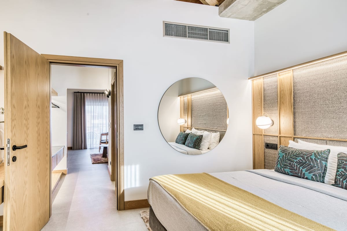 Una habitación de hotel moderna con una cama grande, un espejo redondo, un cabecero de madera y una puerta visible que da a otra habitación.