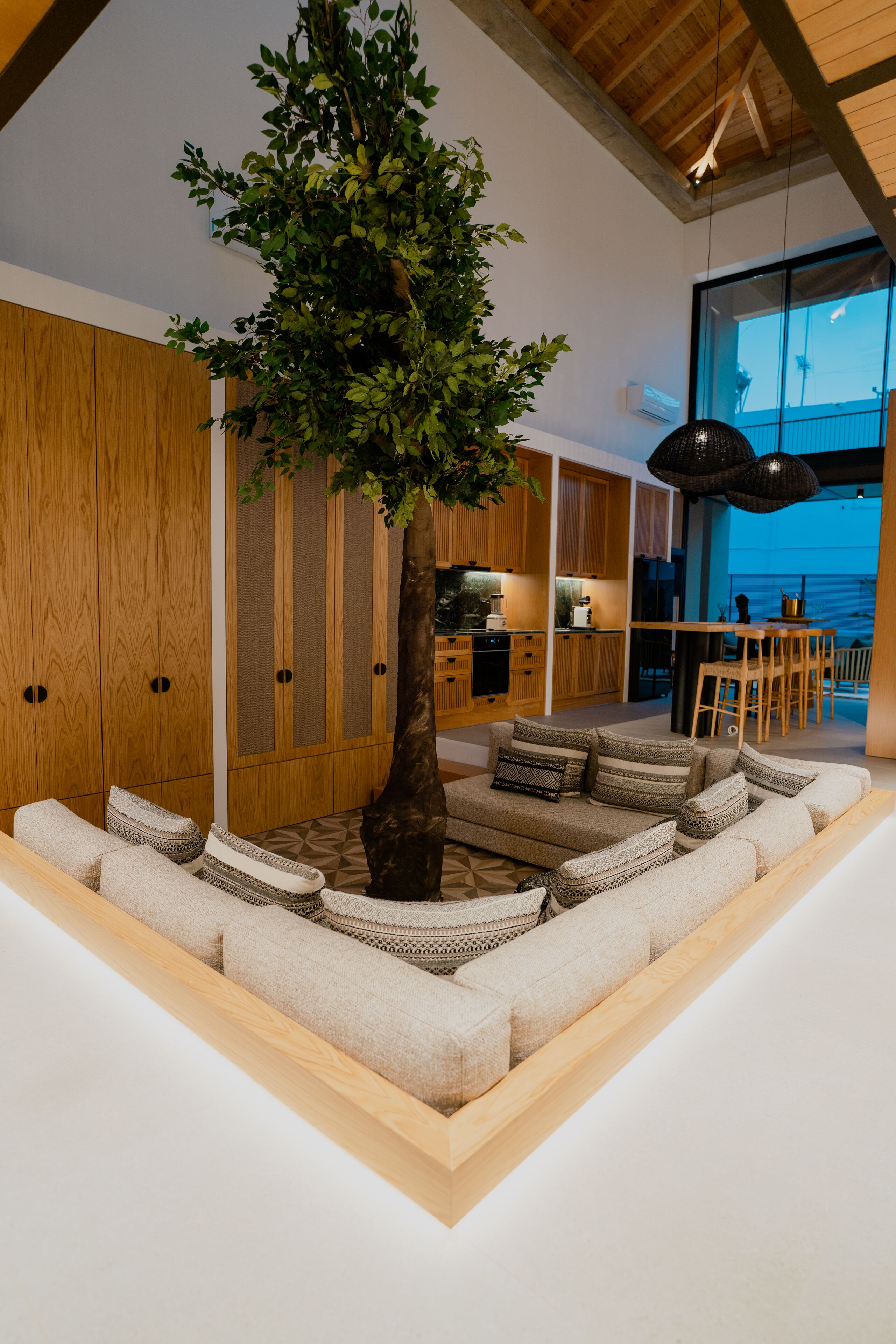 A modern living room with a large tree, wooden cabinets, a sofa with patterned cushions, and a dining area with a table and chairs.