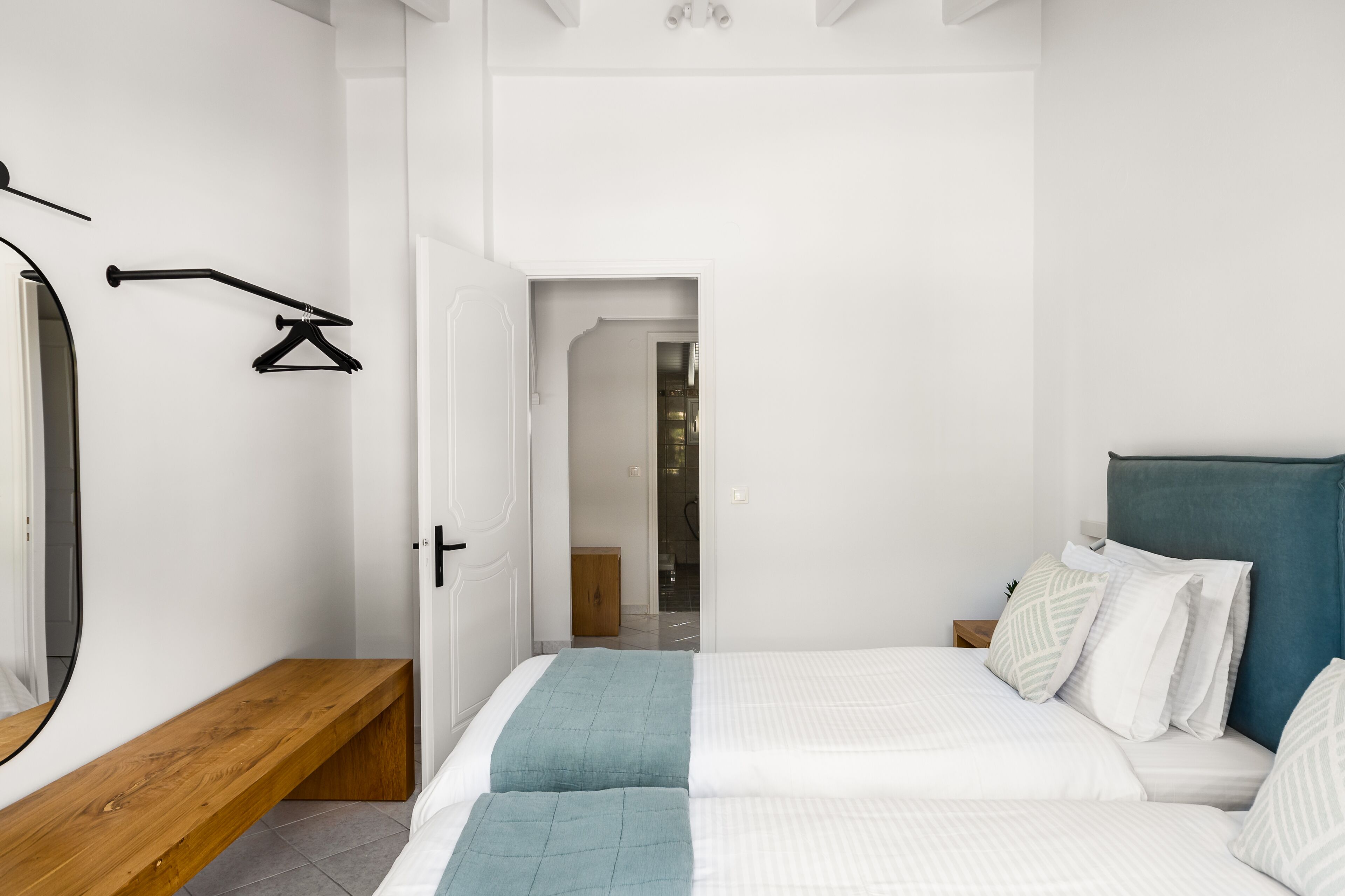 A modern bedroom with a large bed, a wooden bench, a mirror, and a door leading to another room.