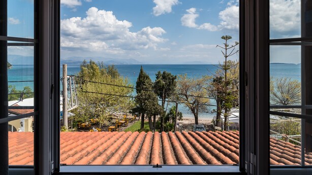 A view from a window overlooking a beach with a clear sky, trees, and a tiled roof. - The Seaview Corner Apartments by Konnect, Ipsos (Corfu)