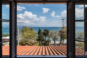 Une vue depuis une fenêtre donnant sur une plage, avec un ciel dégagé, des arbres et un toit recouvert de tuiles.