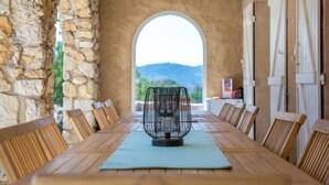 Een lange houten tafel met een metalen decorstuk in het midden, omringd door houten stoelen, in een kamer met stenen muren en een rondboogvenster.