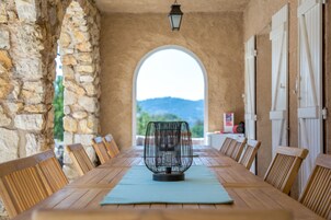 Una larga mesa de madera con un centro de mesa metálico, rodeada de sillas de madera, en una habitación con paredes de piedra y un ventanal arqueado.