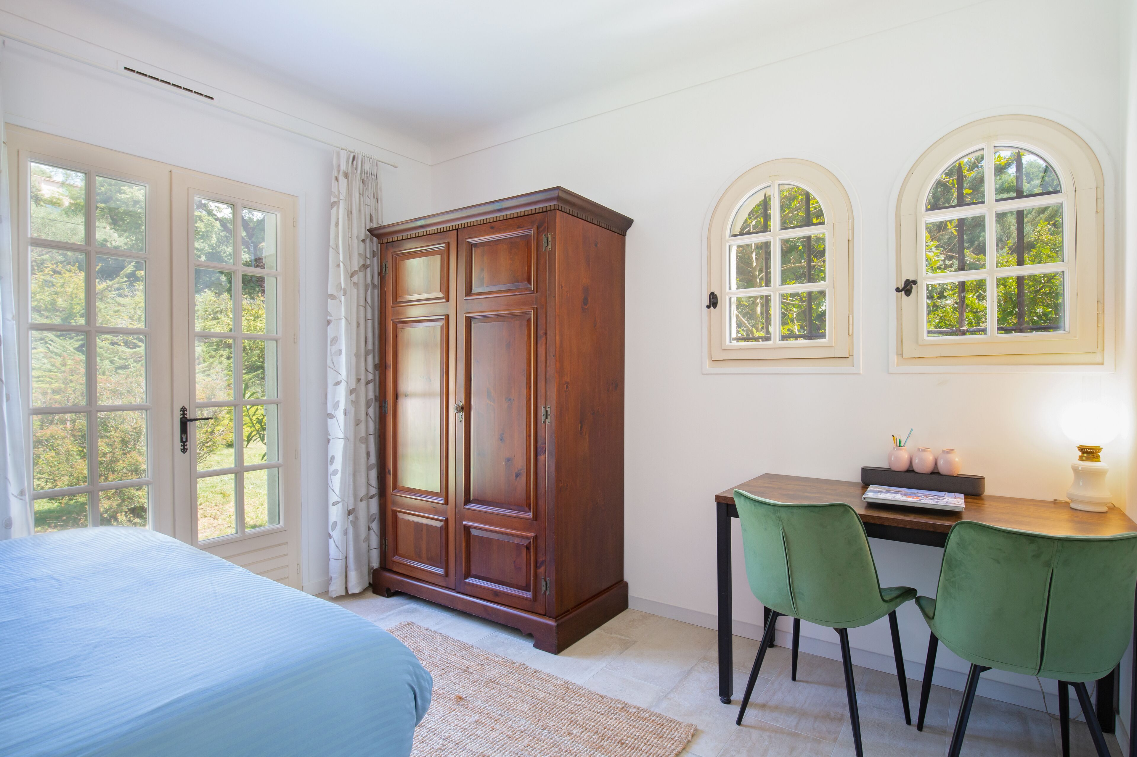 A bedroom with a bed, a wooden wardrobe, a desk with two chairs, and two arched windows.