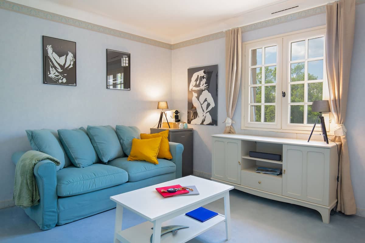 Ein Wohnzimmer mit einem blauen Sofa, einem weißen Couchtisch und einem weißen Schrank. An der Wand hängen zwei gerahmte Bilder, auf dem Schrank steht eine Lampe und es gibt ein Fenster mit Vorhängen.