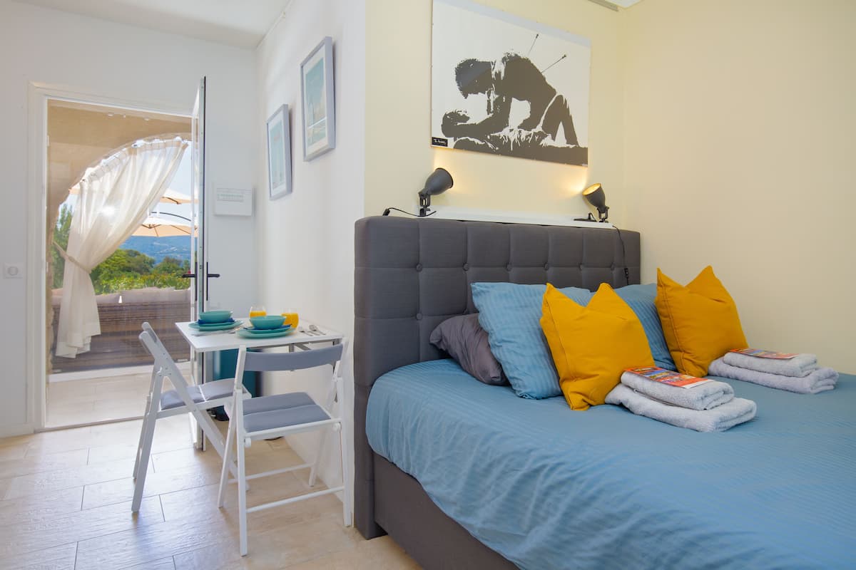 Ein Schlafzimmer mit Bett, Kissen, Nachttisch, Stuhl und Blick durch eine geöffnete Tür.