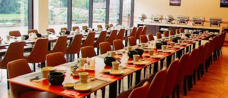 A large dining hall with long tables set for a meal, chairs arranged around each table, and a view of the outdoors through large windows.