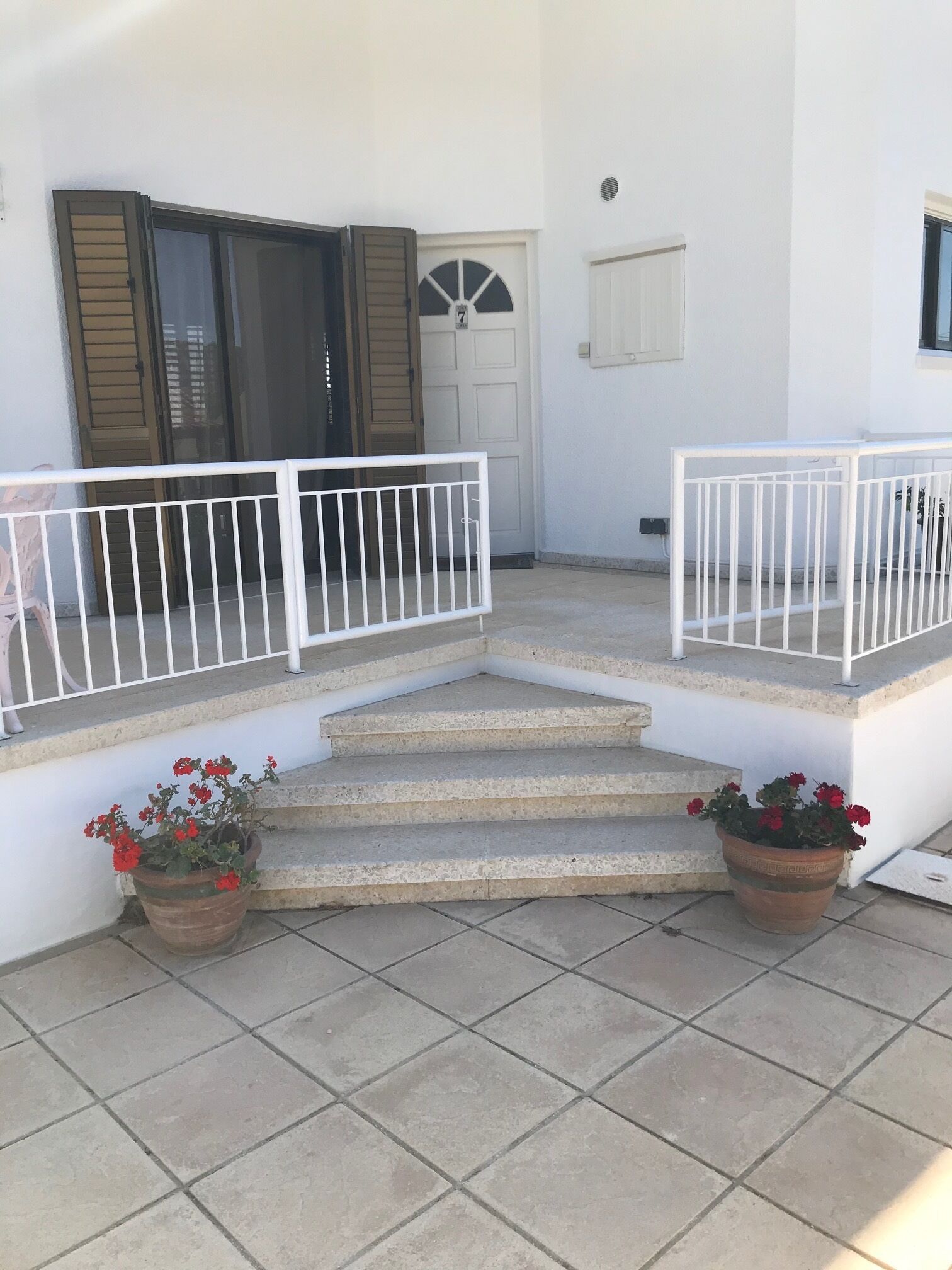 Un bâtiment blanc avec un escalier en béton menant à une porte dotée d’une fenêtre. De part et d’autre de l’escalier, on trouve deux plantes en pot ornées de fleurs rouges.