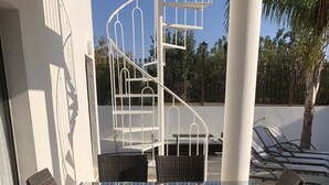 Een tafel met glazen blad en rieten stoelen, een witte wenteltrap en een palmboom op de achtergrond.