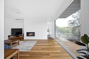 A modern living room with a fireplace, a television, and a large window offering a view of the outdoors. - Sunset on Moonah. (Fingal)