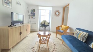 Een woonkamer met een televisie op een houten kast, een rieten stoel met een gele kussen, een blauwe bank en een geweven vloerkleed.