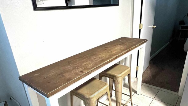 Una cocina con una larga mesa de madera y dos taburetes, una pared blanca y una puerta que da a otra habitación.