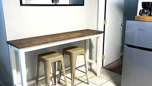 Una pequeña cocina americana con un refrigerador blanco, una barra de madera, dos taburetes y un cuadro enmarcado sobre la barra.