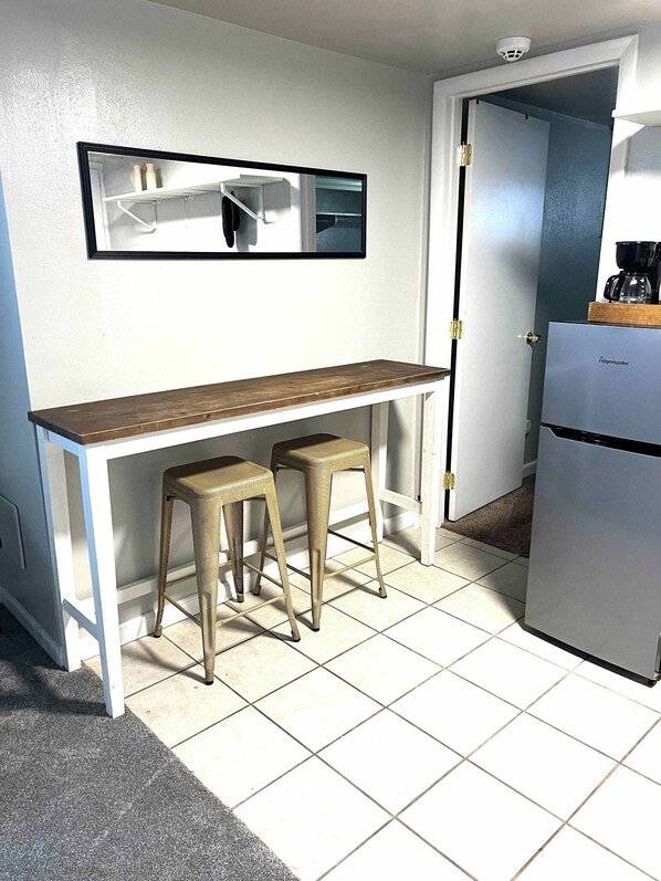 A small kitchenette with a white fridge, a wooden bar counter, two stools, and a framed picture above the counter.