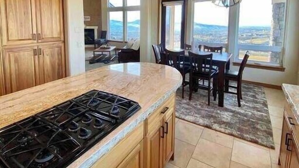 Una cocina moderna con una isla grande, armarios de madera y una estufa a gas. Hay un comedor con una mesa y sillas, y ventanas amplias que ofrecen vistas del paisaje.