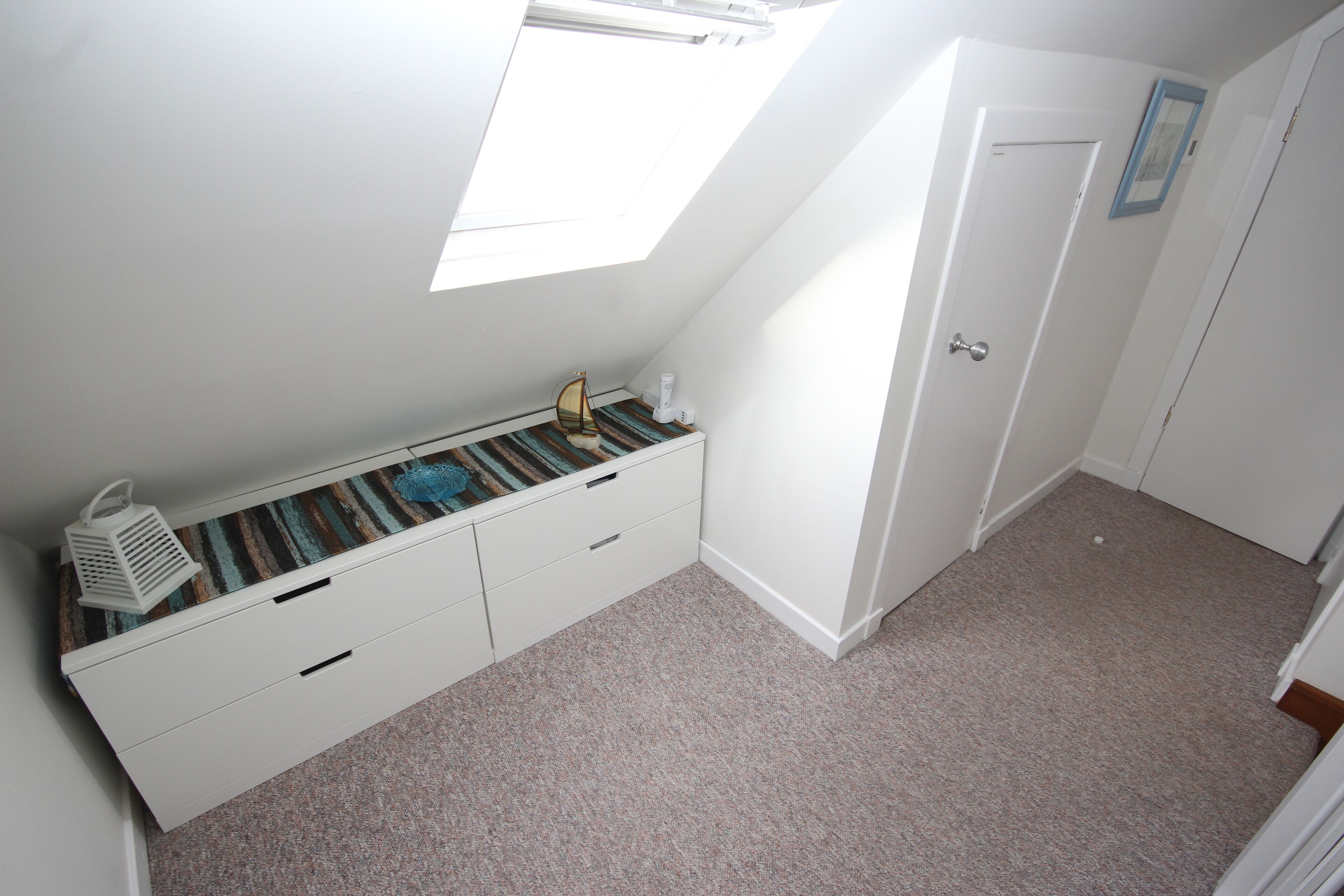 A bedroom with a sloped ceiling, a window, a white door, and a small bedside table with a lamp.