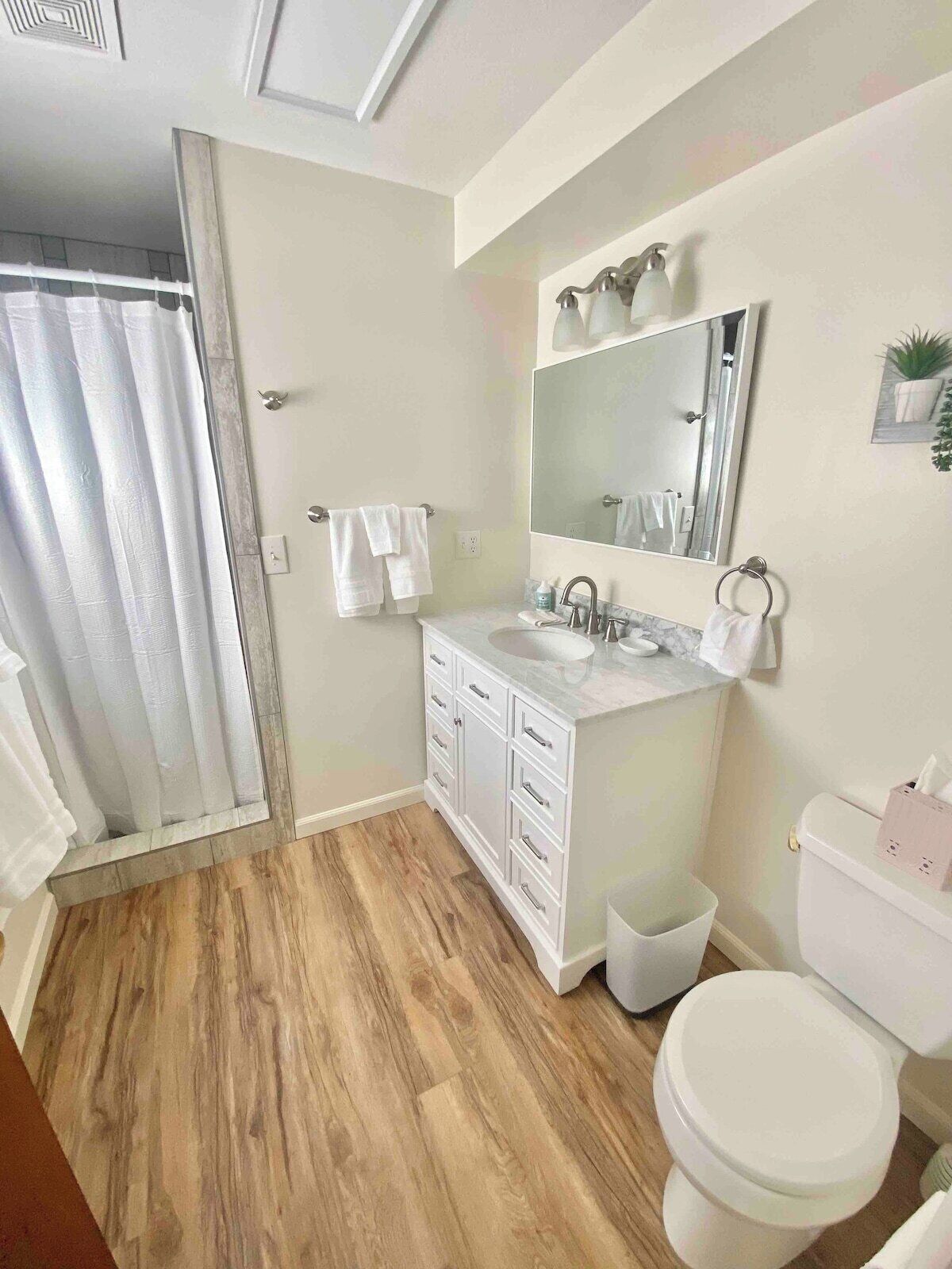 A bathroom with a wooden floor, white cabinets, a sink, a toilet, and a shower curtain.