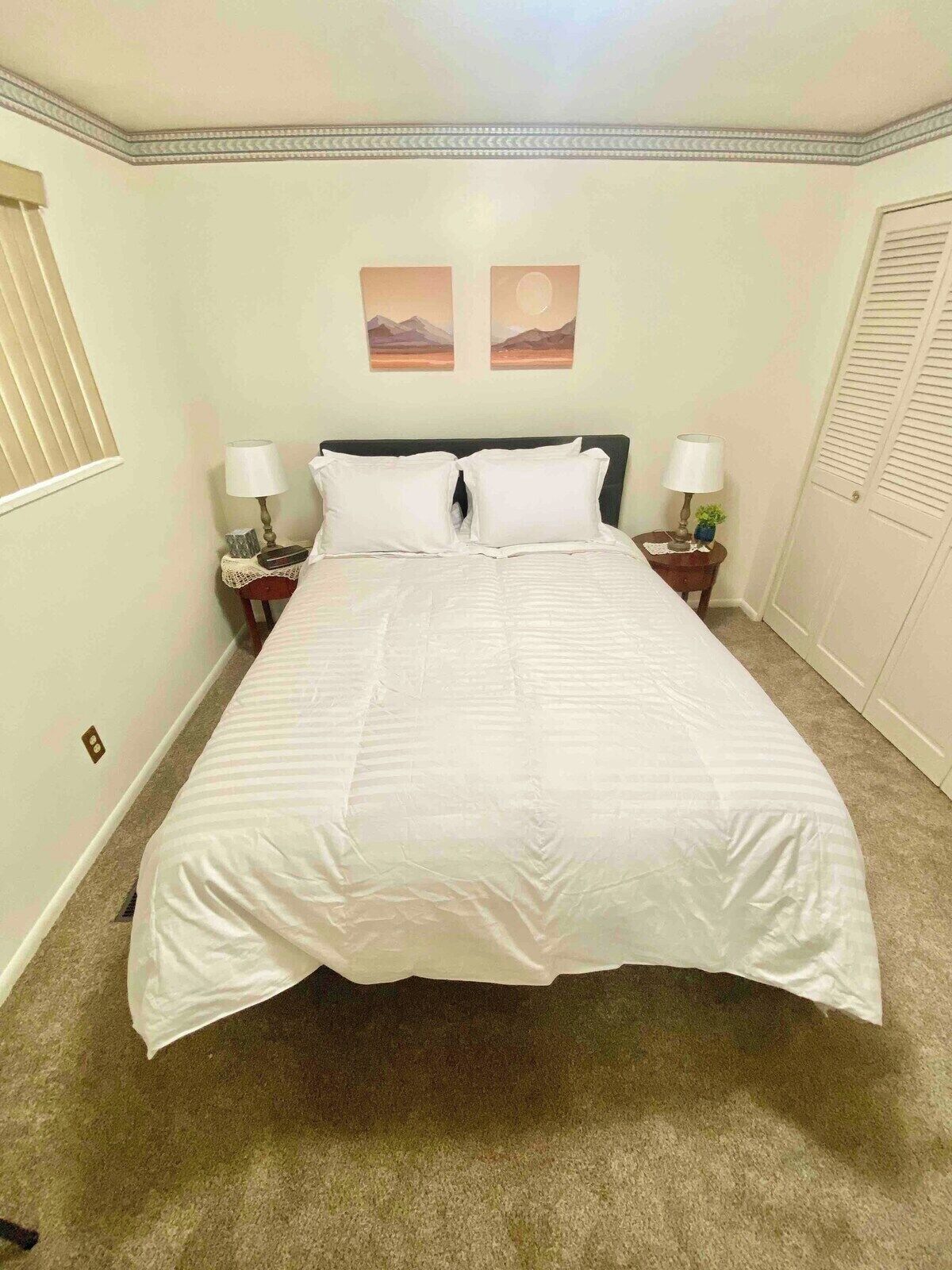 A neatly made bed in a hotel room with a plain wall, two bedside tables with lamps, and a window with blinds.