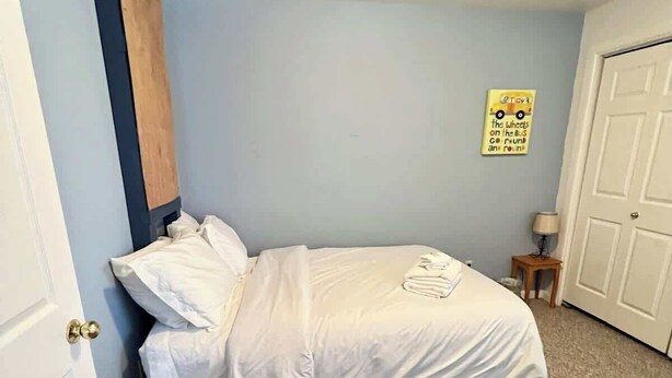 A small, neatly made bed in a room with a blue wall, a wooden headboard, a door, and a small table with a lamp. - Unique Home Near Temple and Parks, great for families! (Pocatello)