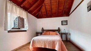 Une chambre avec un plafond en bois, un lit recouvert d’une couvre-lit à motifs, une table de chevet, une fenêtre avec des rideaux et un tableau encadré accroché au mur.