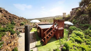 A wooden deck with a table and chairs, surrounded by greenery and a stone wall. - Vacation Home "Casa Rural Romance" with Sea View, Jacuzzi & Wi-Fi (Icod de los vinos)