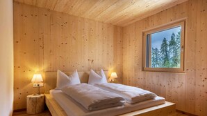 Puinen mökkimäinen huone, jossa on sänky, kaksi yöpöytävalaisinta ja ikkuna, josta avautuu näkymä metsään.