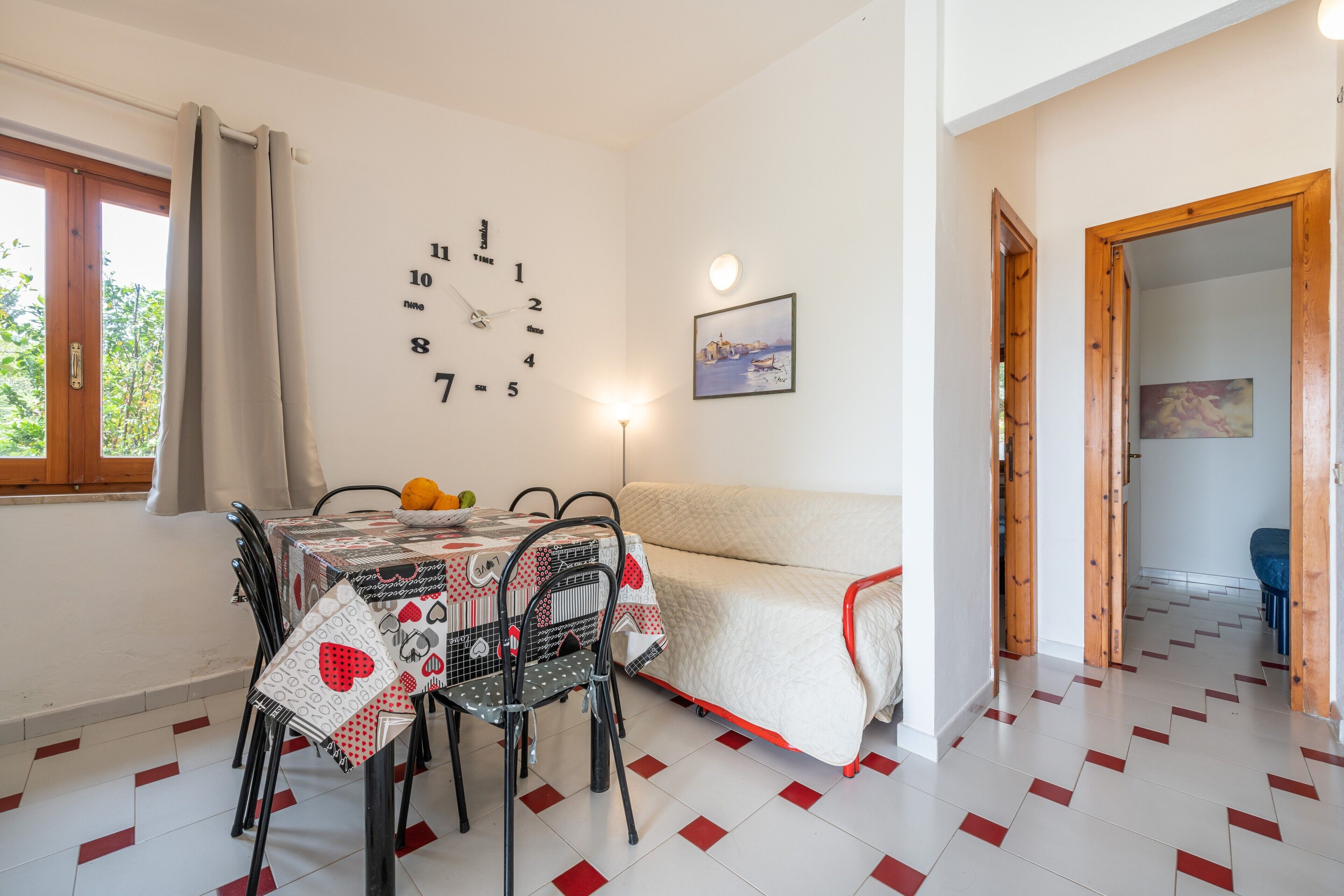 Uma sala de jantar com uma mesa posta para quatro pessoas, um sofá, um relógio de parede e um quadro.