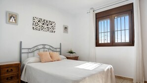 Ein Schlafzimmer mit Bett, Nachttisch, Kommode, einem Fenster mit Vorhängen und Wandbildern.