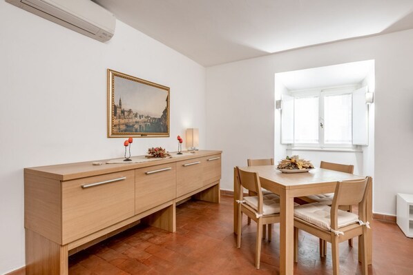 A dining area with a wooden table and chairs, a cabinet with drawers, a framed picture on the wall, and a window with white shutters.