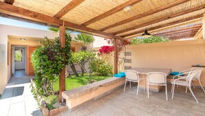 Un patio exterior con una pérgola de madera, un banco de piedra y sillas de metal blanco.