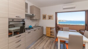 Cozinha moderna com forno, fogão e pia, área de jantar com mesa e cadeiras, e vista para o mar por uma grande janela.