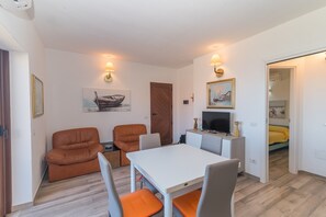 Ein Zimmer mit einem weißen Tisch, zwei orangefarbenen Stühlen, einer braunen Tür und einem Fernsehgerät auf einem kleinen weißen Schrank.