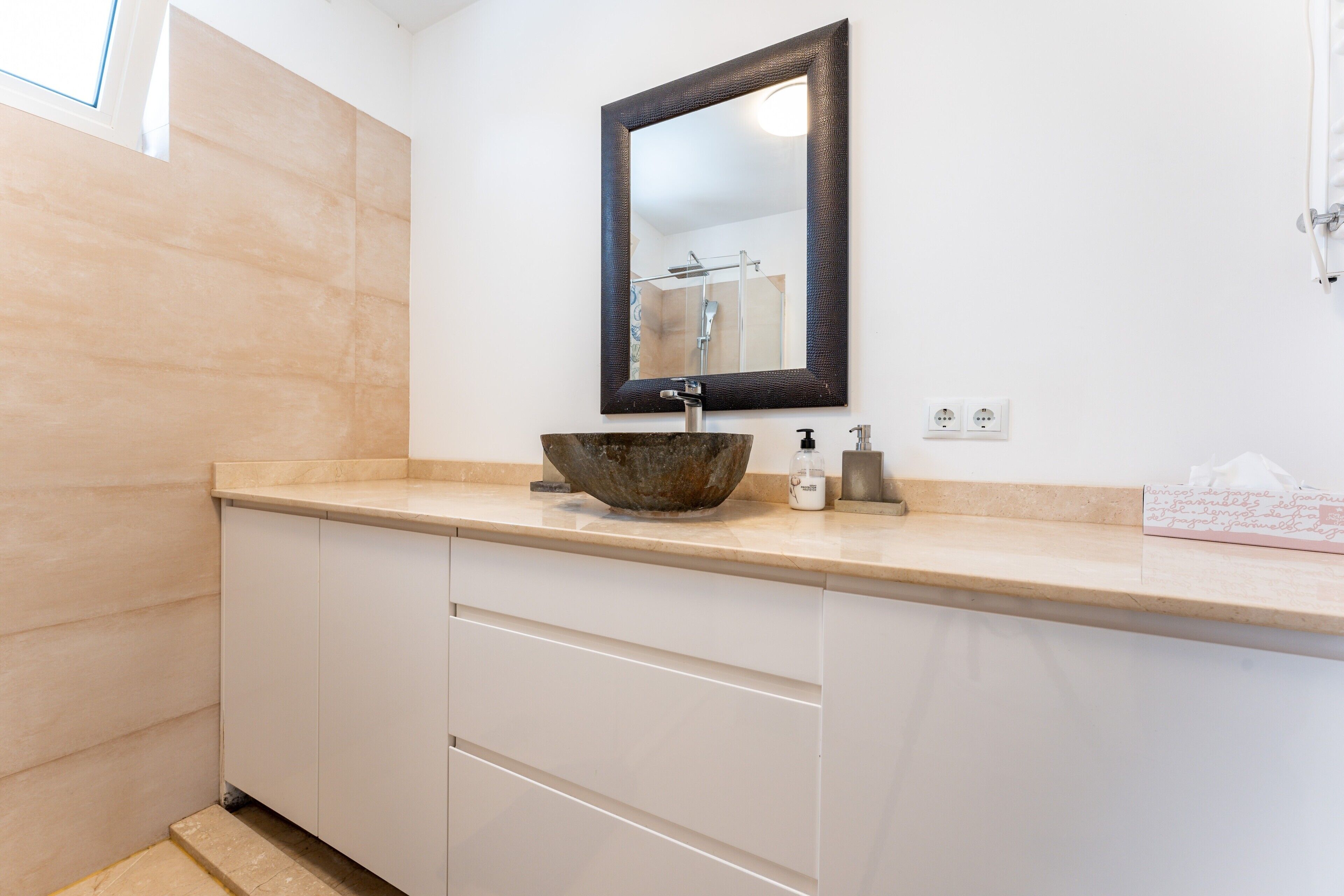 A bathroom with a beige countertop, a large mirror, a vessel sink, a soap dispenser, and a towel rack.
