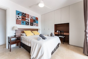 Une chambre à coucher comprenant un lit, des tables de chevet, un bureau, une chaise, un ventilateur de plafond et une œuvre d’art murale.