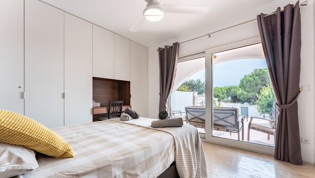 A bedroom with a bed, a desk, a chair, a window with curtains, and a view of the outdoors.