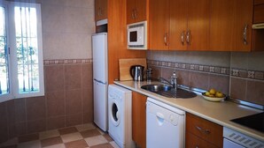 A compact kitchen with wooden cabinets, a built-in microwave, a washing machine, a dishwasher, and a sink with a bowl of fruit on the counter.