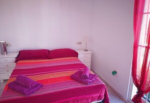 A bed with a striped bedspread, two pillows, a nightstand with a lamp, and a window with red curtains.