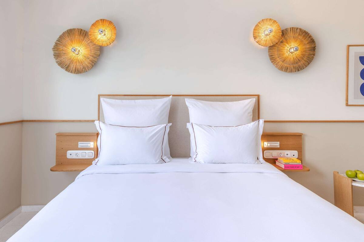 A neatly made bed with white linens, two bedside tables with lamps, and a small shelf with books and apples.