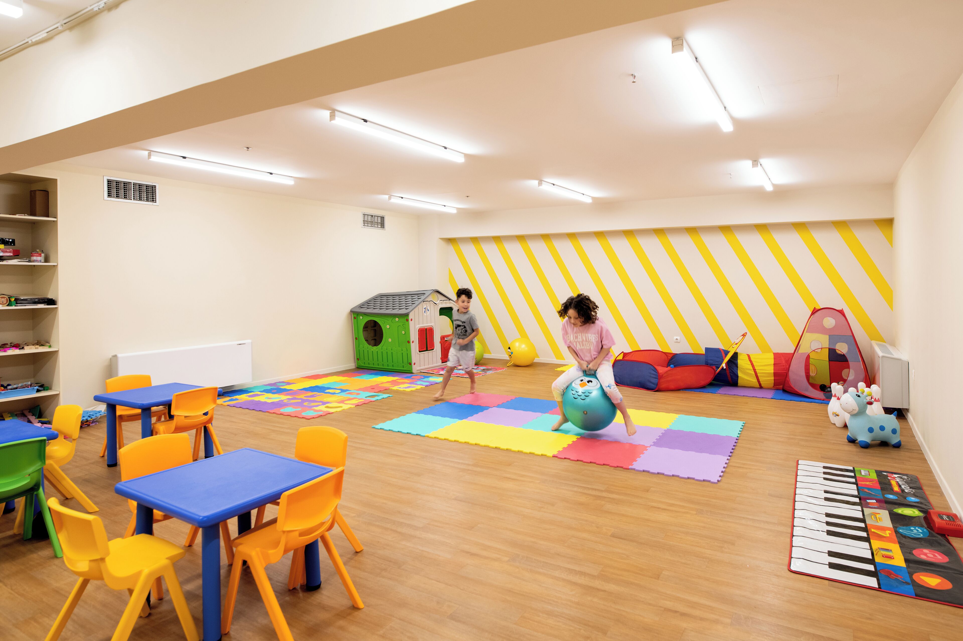 A brightly lit room with wooden floors, colorful play mats, and various children's toys.