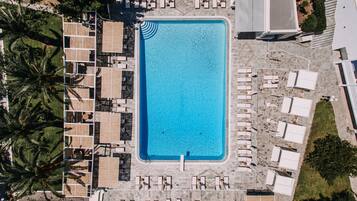 An aerial view of a swimming pool surrounded by lounge chairs and umbrellas, with palm trees and a tiled deck.