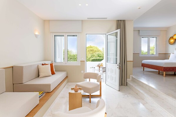 A modern hotel room with a sofa, armchair, and a bed. There are large windows and a view of greenery outside.