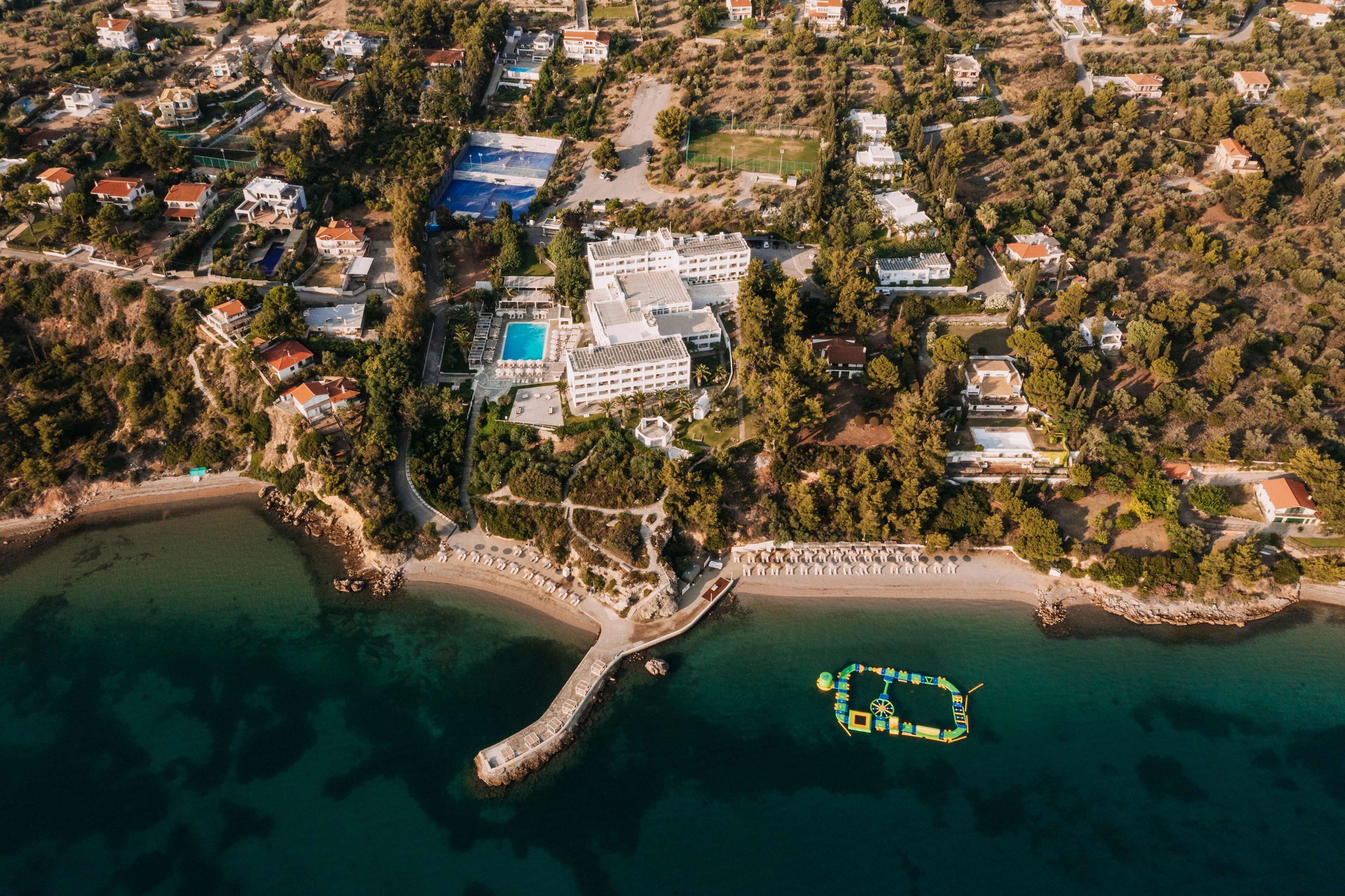 An aerial view of a coastal resort with a central building surrounded by villas, a beach, and a pier.